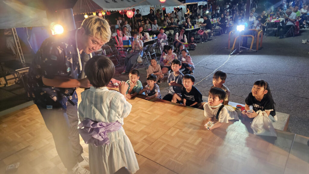 納涼祭のマジックショーで浴衣姿の子どもと共演し、舞台前に集まった子どもたちが興味津々で見入る様子