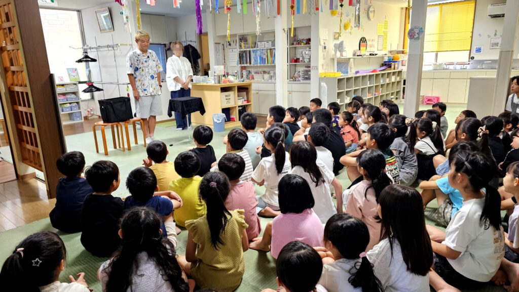 学童イベント マジックショーの開演前、マジシャンと理事長が前に立ち、子どもたちが熱心に見つめている様子