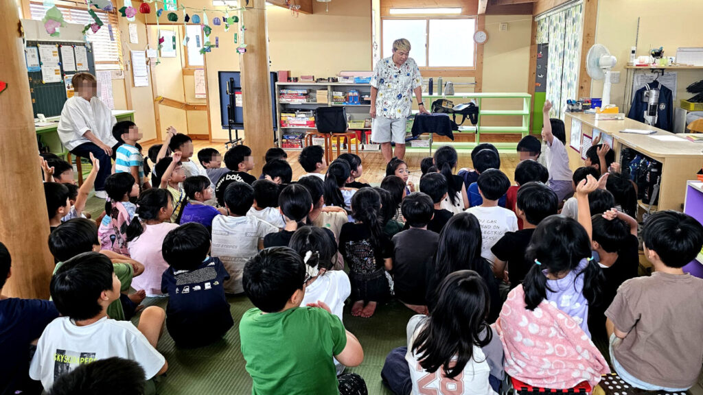 学童イベント マジックショーで、マジシャンが子どもたちに質問を投げかけ、複数の子が手を挙げている様子