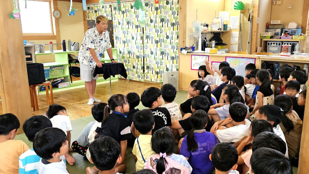 学童イベント マジックショーで、マジシャンがテーブルを使った演目を披露し、子どもたちが集中して見守っている様子