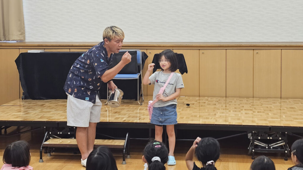 ステージ上で古典マジック『マイザーズドリーム』を演じるマジシャンと、サポート役として前に出た女の子。観客の子どもたちが興味津々で見つめている様子。