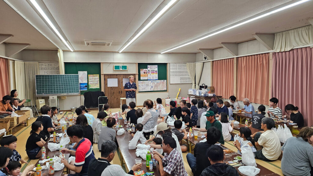 高岡市の納涼祭で行われたマジックショー。室内会場で演者が立ち、食事を楽しむ地域の人々が笑顔でステージを見守っている様子。