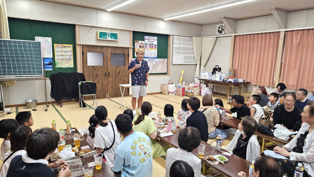 高岡市の納涼祭で行われたマジックショー。室内会場で演者が立ち、食事を楽しむ地域の人々が笑顔でステージを見守っている様子。