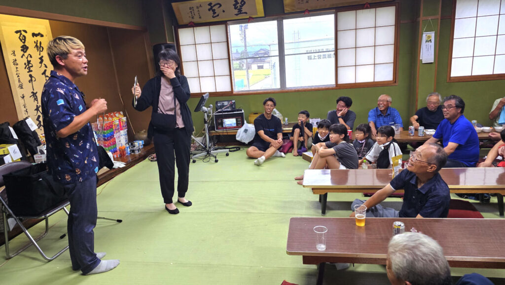 地域納涼祭の室内会場でのマジックショー。観客の笑顔と拍手の中、参加者がステージでマジック体験に挑戦