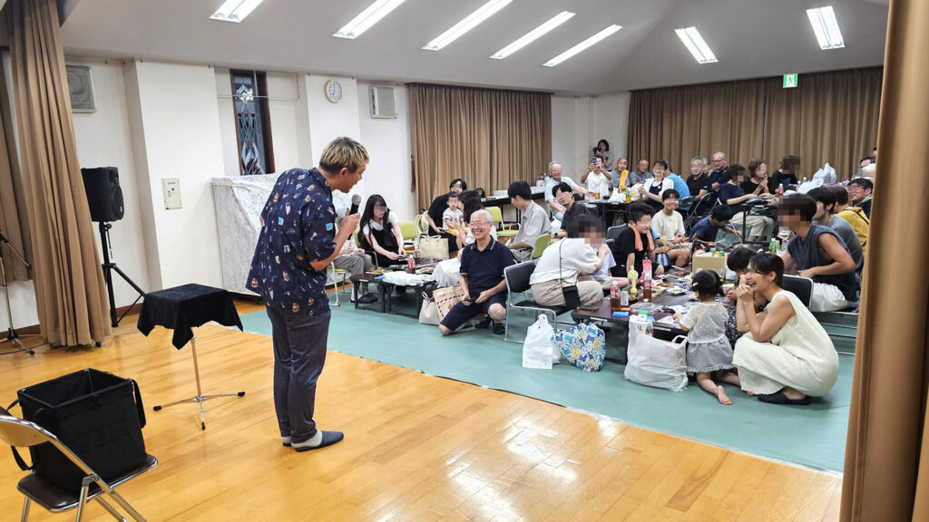とやまアーティストマッチング事業を活用した町内の納涼祭で、マジシャンがマイクを手にマジックショーを披露し、会場の子どもから大人までが笑顔で見入っている様子