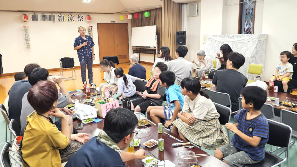 とやまアーティストマッチング事業を活用した町内の納涼祭で、マジシャンがマジックショーを披露し、子どもから大人まで地域の参加者がテーブルで飲食を楽しみながら見入っている様子