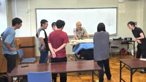 マジシャンのコンプレッサーさんを囲み、福井県立武生高等学校の生徒と先生が文化祭のイリュージョンショーの練習をしている様子。教室の中で、皆さん笑顔で楽しそうに打ち合わせをしています。