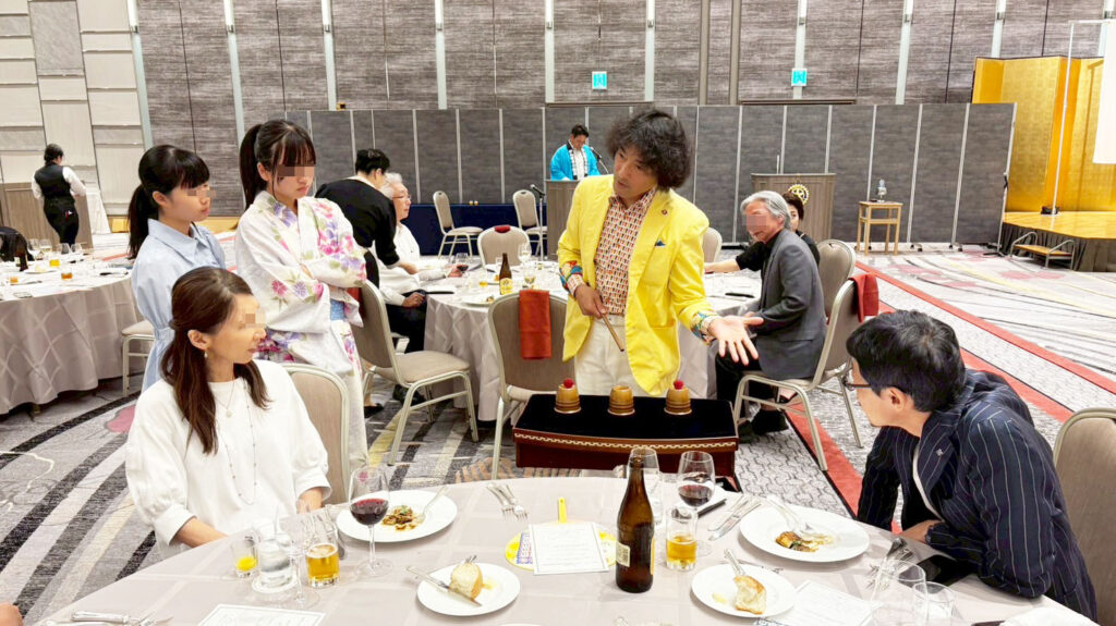 ホテル日航金沢での金沢ロータリークラブのパーティーにて、アシスタントのともやんがテーブルマジックを披露し、観客が囲んで楽しんでいる様子。