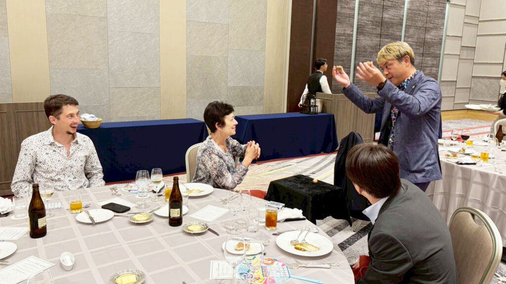 金沢ロータリークラブのパーティーで、マジシャンがテーブルマジックを披露し、外国人ゲストを含む観客が驚きと笑顔で楽しんでいる様子。