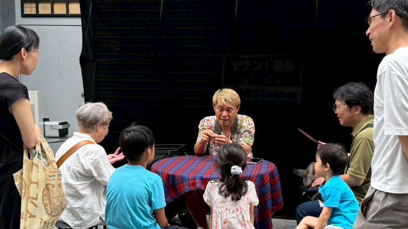 富山ストリートパフォーマンスでマジシャンが観客の子どもたちに囲まれ、テーブルマジックを真剣に披露している場面