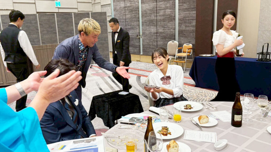 金沢ロータリークラブのパーティーで、マジシャンがテーブルマジックを披露し、観客がカードを手に笑顔で楽しんでいる様子。