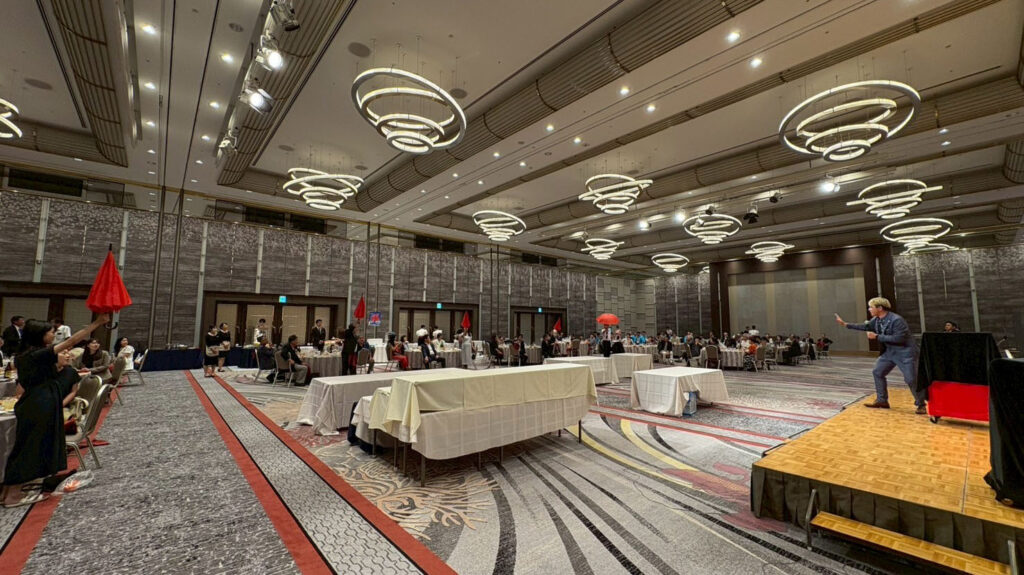 ホテル日航金沢の大広間で行われた金沢ロータリークラブのパーティーにて、ステージ上のマジシャンと観客が赤い旗を使ったマジックに参加している様子。