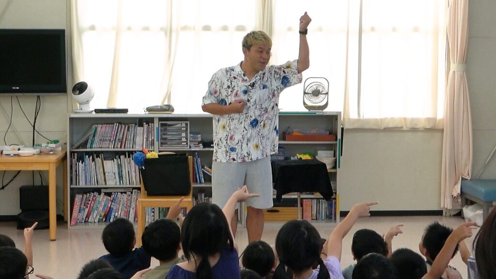 高岡市の学童保育でのマジックショー。コンプレッサーが子どもたちに問いかけ、元気に手を挙げている場面。