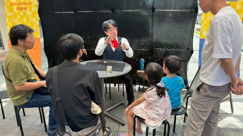 富山ストリートパフォーマンスでベテランマジシャン　まぁくん　がテーブルマジックを披露し、観客の子どもたちや大人が集中して見入っている場面