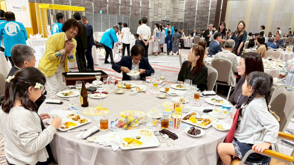 金沢ロータリークラブのパーティーで、アシスタントのともやんがテーブルマジックを披露し、観客の家族連れが笑顔で楽しんでいる様子。