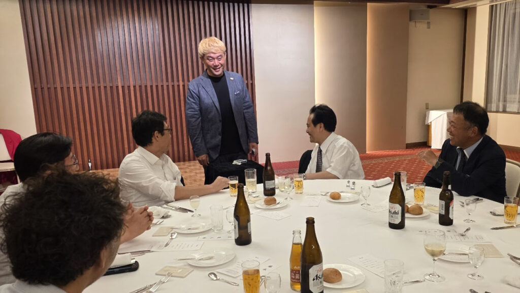 企業パーティーの懇親会で、円卓を囲む参加者にテーブルマジックを披露するマジシャン・コンプレッサー。