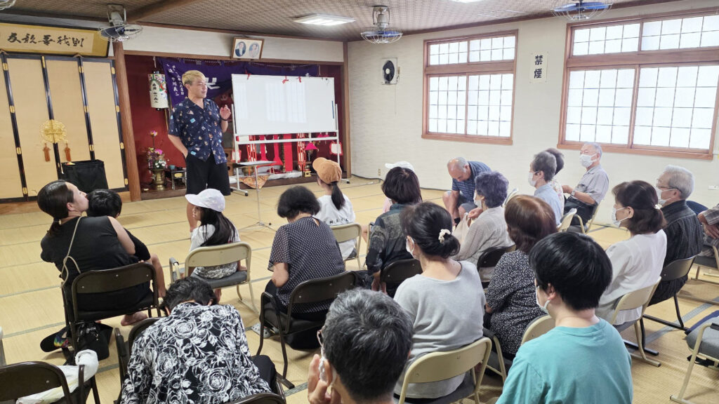 【出演レポート】高岡の地蔵祭りが教えてくれた、地元「福岡町 つくりもんまつり」の知らなかった原点 1 公民館のような和室で、マジシャン・コンプレッサーが集まった観客に向かって話している様子。観客は老若男女で、椅子に座って真剣な表情でショーを見ている。