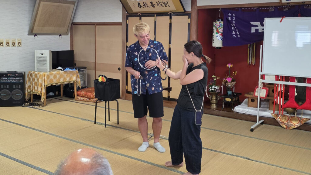 【出演レポート】高岡の地蔵祭りが教えてくれた、地元「福岡町 つくりもんまつり」の知らなかった原点 2 畳の部屋で行われているマジックショーの様子。金髪のマジシャン・コンプレッサーが観客の女性にリングを渡しており、女性は驚きの表情で口元に手をやっている。
