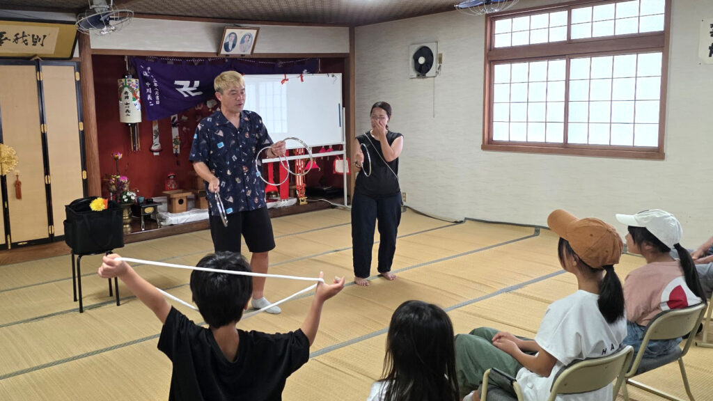 【出演レポート】高岡の地蔵祭りが教えてくれた、地元「福岡町 つくりもんまつり」の知らなかった原点 3 マジシャン・コンプレッサーが観客参加型のマジックを披露している。一人の少年が白いロープの両端を持って立ち上がり、その後ろでは女性の観客が驚いた表情を見せている。