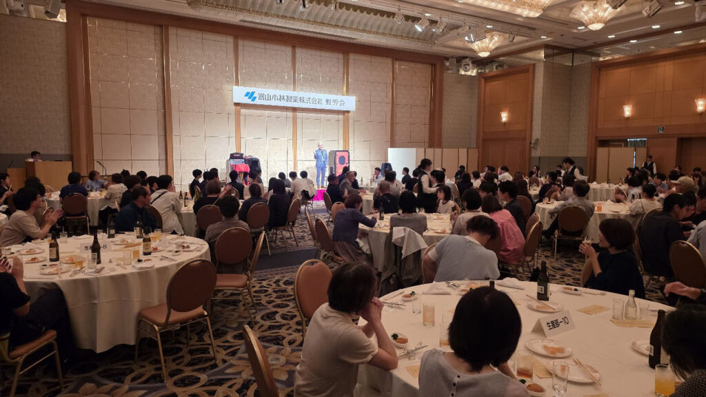 富山でのパーティー余興で、傘を次々と出現させる華やかなマジックを演じるコンプレッサー。大人数のゲストがその見事なテクニックに見入っている。