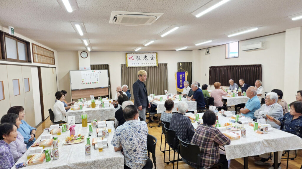 敬老会のイベント会場をマジックで盛り上げるマジシャン・コンプレッサー。富山県高岡市の公民館に集まった大勢の参加者が、テーブルを囲んで楽しそうにショーを見ている様子。