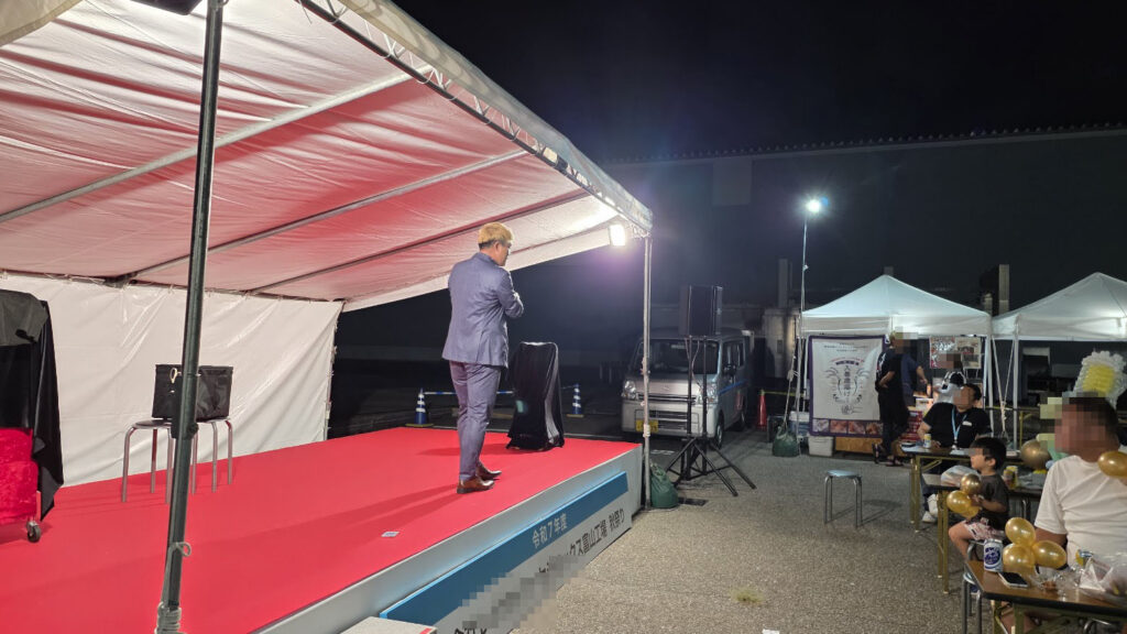 企業イベントのステージに立つマジシャン・コンプレッサーの後ろ姿。赤いカーペットが敷かれたステージ上から、観客席や屋台のテントが見える。