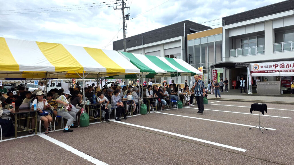 中田かかし祭の屋外会場でマジックを披露するコンプレッサーのワイド写真。緑と黄色のテントの下に多くの観客がいる。