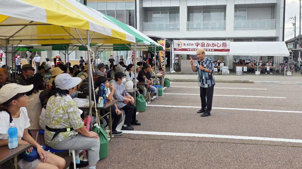 中田かかし祭の会場で、テントの下に座る観客を前にマイクを持って話すマジシャン・コンプレッサー。