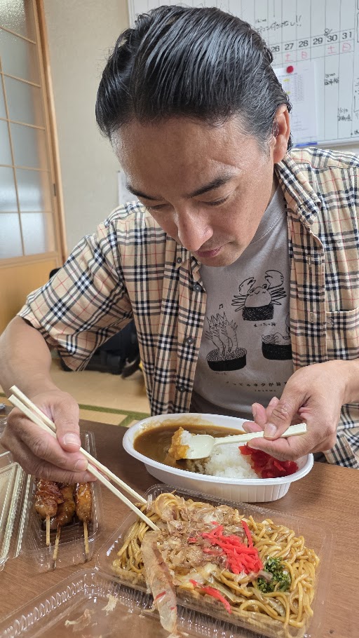 射水市中村自治会の秋祭りで提供された昼食を食べるマジシャン・コンプレッサーのアシスタント「ともｙはん」。カレーライス、焼きそば、焼き鳥などボリューム満点のメニューが並ぶ。