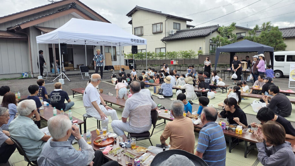 射水市中村自治会の秋祭りの全景。子どもから高齢者まで多くの地域住民が集まり、ござの上で飲食を楽しみながらマジックショーを観覧している様子。