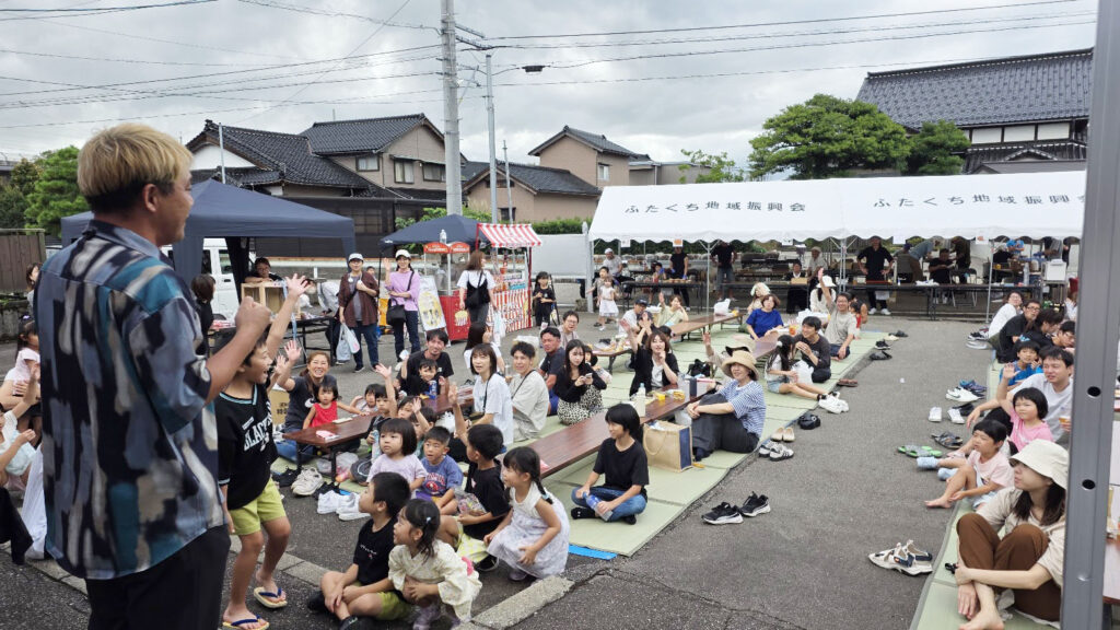 マジックショーに沸く射水市中村自治会秋祭りの観客。最前列に座る子どもたちが笑顔でショーに見入っており、会場の一体感が伝わる一枚。