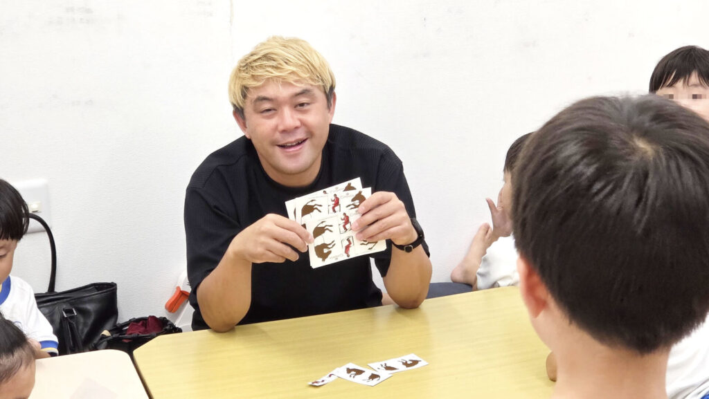 マジシャン・コンプレッサーが子どもたちに囲まれ、笑顔でトリックアートのカードを見せながら説明している。