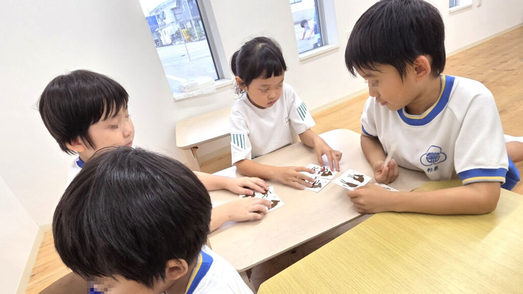 明るい室内で、4人の子どもたちがテーブルを囲み、それぞれがトリックアートのカードを熱心に見つめ、考えている。