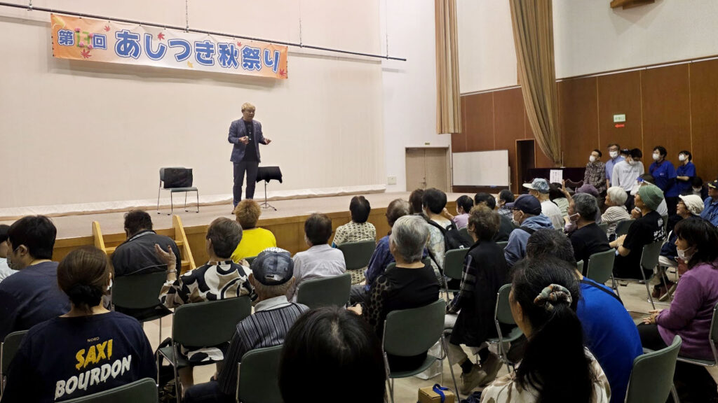 ステージ上でマジックを披露するマジシャン・コンプレッサー。「第13回 あしつき秋祭り」の横断幕が掲げられている。