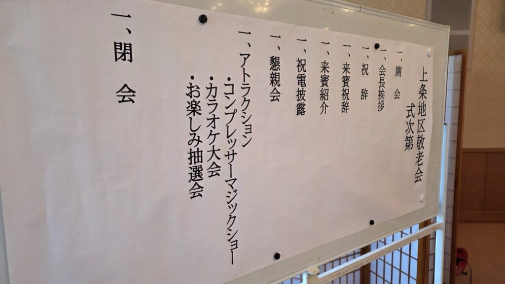 ホワイトボードに貼られた「上条地区敬老会 式次第」。プログラムには「アトラクション」の項目があり、「コンプレッサーマジックショー」の文字が見える。