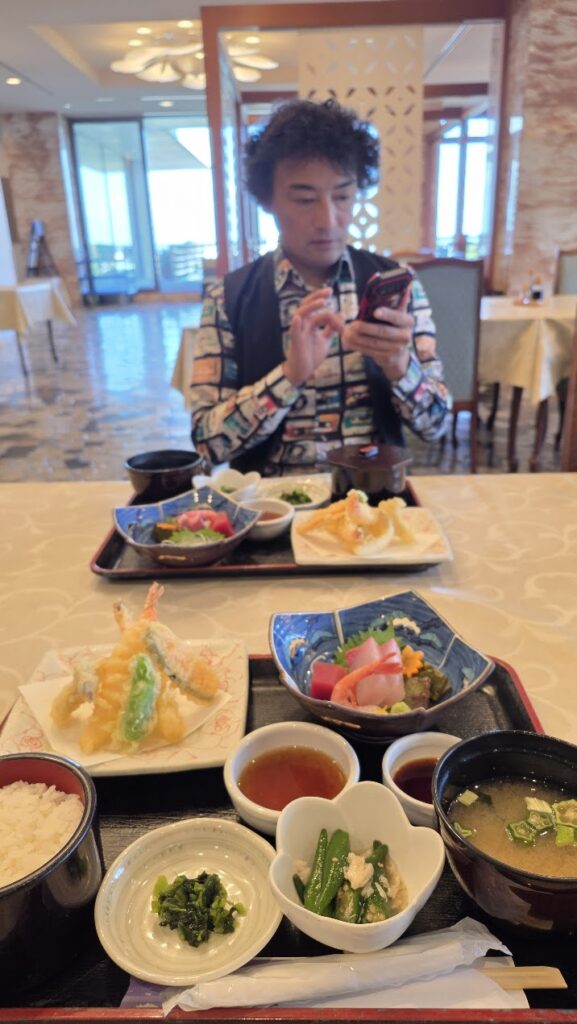 イベント会場のレストランで、天ぷらや刺身が並んだ豪華な和食御膳を前にして、スマートフォンを操作するマジシャン・ともやん