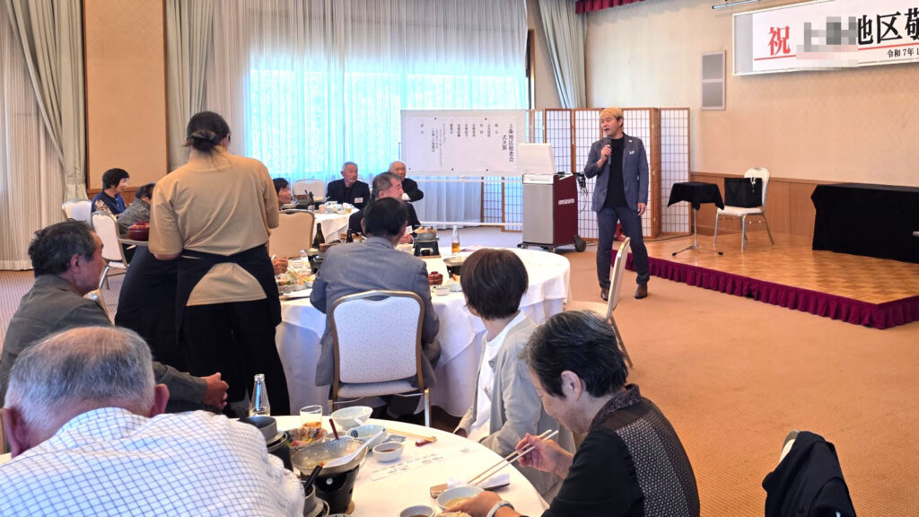 ホテルの宴会場のような広い部屋で開かれている敬老会の様子。ステージに立つマジシャン・コンプレッサーが、円卓を囲んで食事を楽しむお年寄りの観客に向けてパフォーマンスをしている。