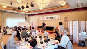 敬老会のステージでマイクを持って話すマジシャン・コンプレッサー。食事をしながらショーを観覧する参加者たちの後ろ姿が見える。