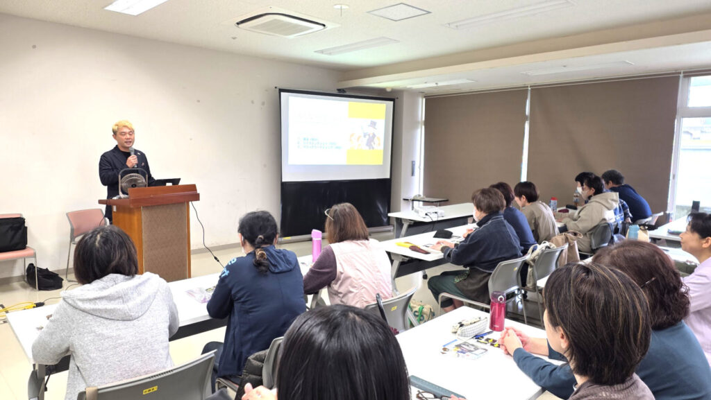 富山県の放課後児童支援員研修会に登壇。マジシャンが伝えたかった「子どもたちと心を通わせる」3つの柱 3 教壇に立ち、マイクを持って受講者に語りかけるマジシャン・コンプレッサー。受講者が真剣な表情で話を聞いている。