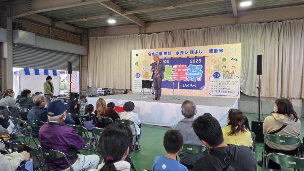 【JAくろべ農業祭】2回公演の熱気!マジシャンが語る「現場の数と成長」の法則 4 JAくろべ農業祭のステージに立つマジシャン・コンプレッサー。客席の最前列に座る子供たちに笑顔で語りかけている様子。