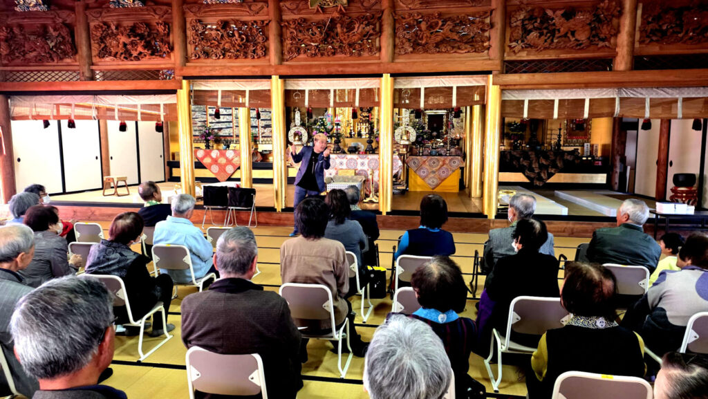 高岡市のお寺の本堂で、コンプレッサーが観客に語りかけ、不思議な現象を見せているステージの様子。