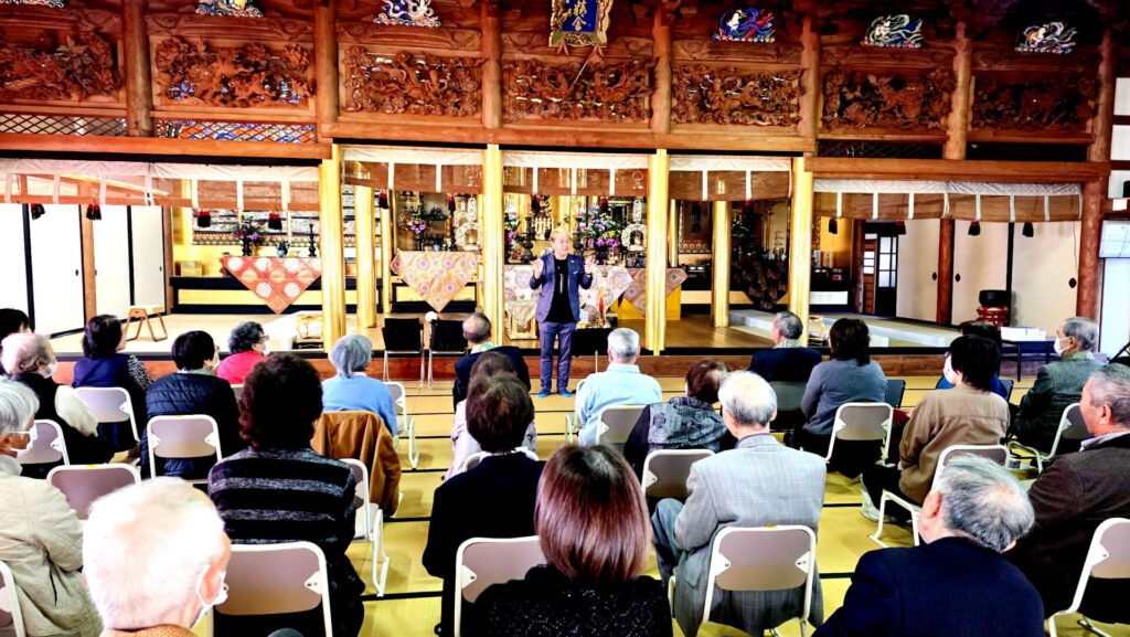 お寺の本堂の畳に座る保育園児と、椅子に座る大人（お年寄り）たち。ステージ中央でコンプレッサーが講演・トークを行っている。