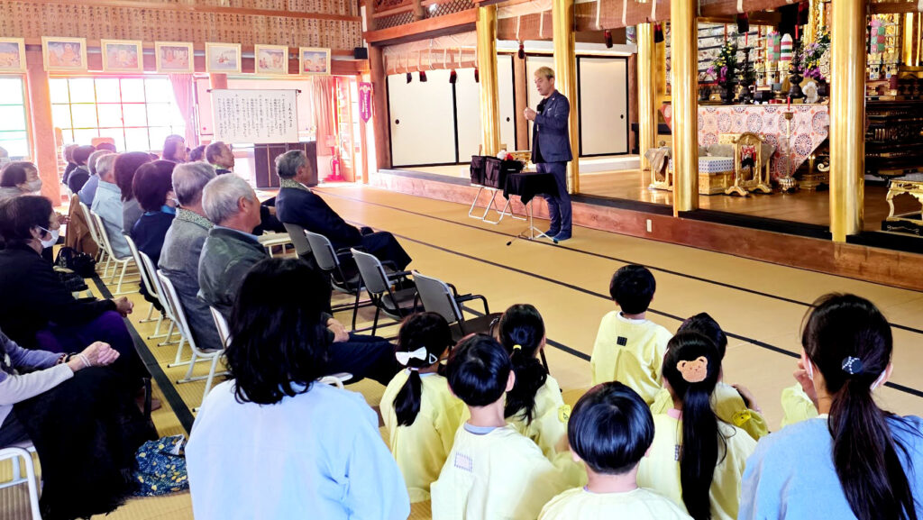 高岡市のお寺の本堂で、コンプレッサーが出演者・お年寄り・園児たちを前にマジックショーを行う様子。