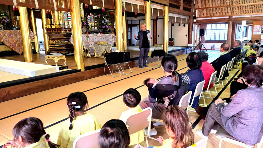 お寺の本堂の格式高い雰囲気の中で、コンプレッサーが聴衆にトーキングマジック（おしゃべりマジック）を披露している様子。