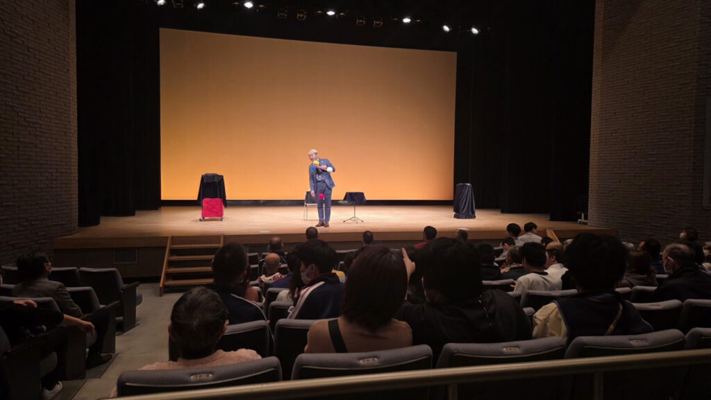 新川文化会館のステージでマジックを披露するコンプレッサーと、集中して見入る魚津市障害者連合会の皆様