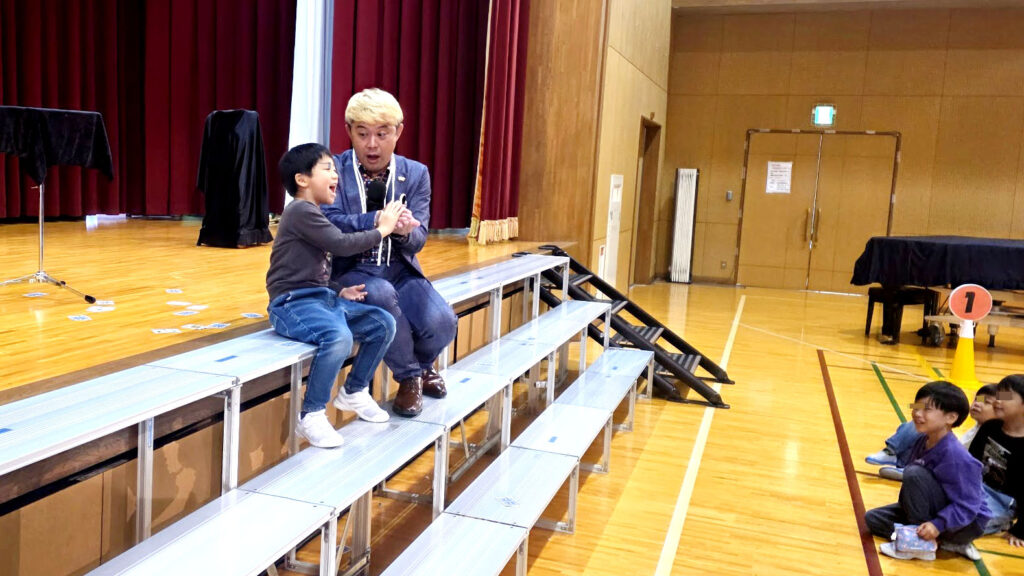 ステージの階段で児童と隣り合って座り目の前でマジックを披露するコンプレッサー