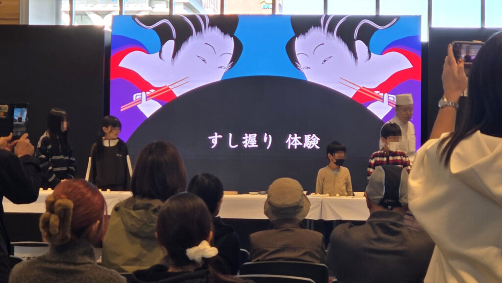 ステージ上で寿司握り体験に参加する子供たちと、それを見守る観客の様子