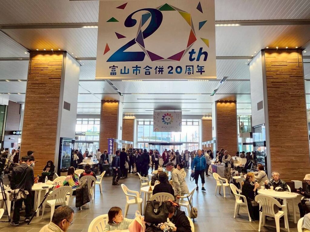 富山市合併20周年のバナーが掲げられた富山駅南北自由通路のイベント会場と、多くの来場者で賑わう様子