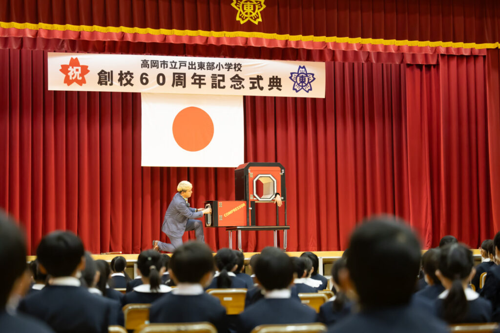 体育館のステージで大掛かりなイリュージョンを披露し、児童を驚かせるマジシャン・コンプレッサー