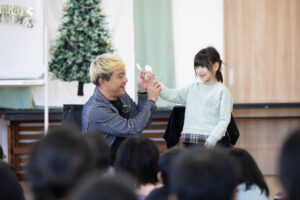 ステージ上で小さな女の子と触れ合いながらマジックを披露するコンプレッサー。女の子の弾けるような笑顔と、優しく寄り添うマジシャンの人間味あふれるコミュニケーションの瞬間。